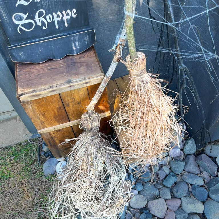 Closeup photo of two handmade witches brooms in a corner in a spooky scene outside with cobwebs. The backdrop is dark gray brick with and there is an old wood crate with a sign on top.