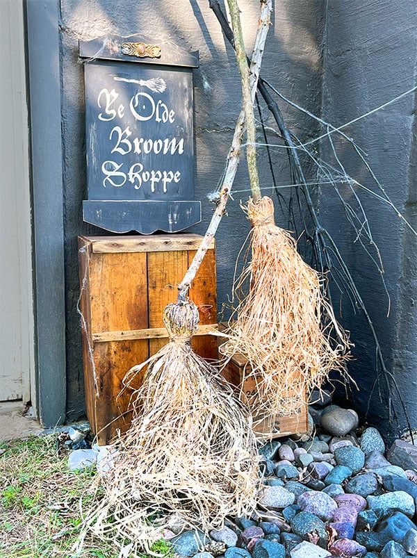 Photo of two handmade witches brooms in a corner in a spooky scene. The backdrop is dark gray and there is an old wood crate with a sign that says Ye Olde Broom Shoppe. The two brooms are in the corner with an eerie tree branch and faux cobwebs. Everything looks like it's been there for a while and the sign is worn and tattered in black with fluted edges top and bottom in off white with a broom illustration over top.