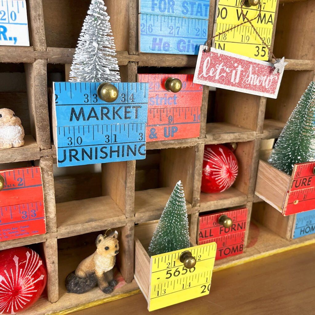 Closeup photo of a yellow sectioned soda crate with twelve tiny drawers made with colorful red, yellow and blue wood yardsticks for the fronts. This soda crate turned display and storage crate is decked out here with tiny Christmas items like resin rabbits and a fox, a teddy bear and vintage red glass ornament orbs filling the holes. Three of the drawers are pulled out to hold bristle brush Christmas trees.
