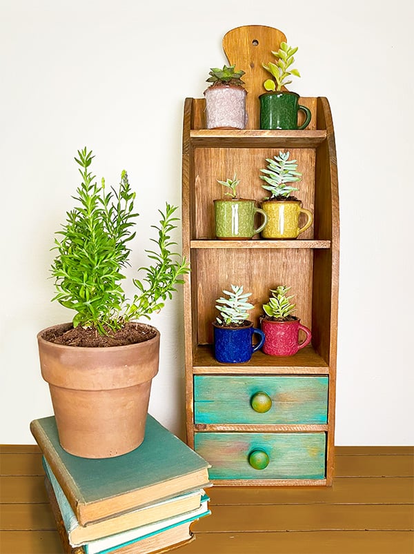 Shown is a pretty vertical wooden wall shelf three tiny shelves and a handle at the top that resembles a wood cutting board handle. The shelf holds six tiny colorful mugs with succulents. A terrecotta planter on a set of aqua faded books. This piece only needed a touch of paint for a little color pop. I painted the two bottom drawers aqua and sanded some of the color away for a more weathered look.