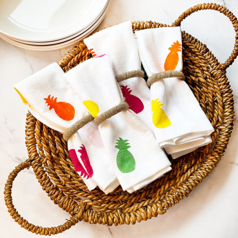 Feature photo for 8 pineapple crafts roundup post that shows an overhead photo of a wicker basket with four white fabric napkins with jute napkin rings in the center that feature colorful fuchsia, spring green, yellow and orange pineapple silhouettes. This is just one of eight easy pineapple crafts in this series to brighten up your home.