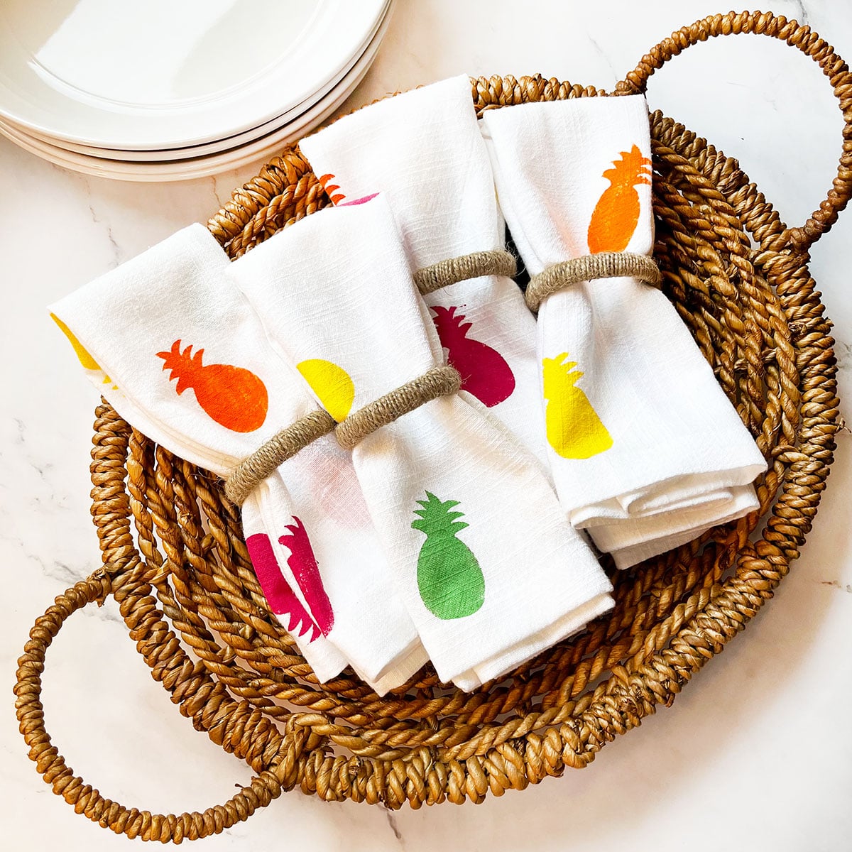 Feature photo for 8 pineapple crafts roundup post that shows an overhead photo of a wicker basket with four white fabric napkins with jute napkin rings in the center that feature colorful fuchsia, spring green, yellow and orange pineapple silhouettes. This is just one of eight easy pineapple crafts in this series to brighten up your home.