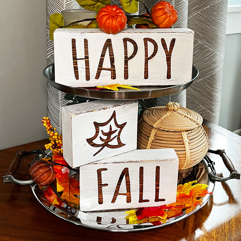 Happy Fall block letters on a tiered tray with fall florals, orange and yellow oak leaves and tiny pumpkin picks and a decorative small wicker basket filling up the space to the back of the tray.