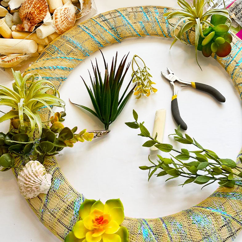 A breezy progress shot and feature photo for 10 fun and easy beachy crafts. shown is a metal wreath form wrapped in sand and aqua tulle with beach florals, succulents, greenery sprigs, shells, and wire snips at the ready.