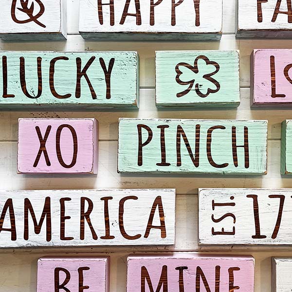 Square feature photo of rows of different stackable word signs made with two by fours. They are for different holidays, pastel painted pink for XO, Be Mine, Lucky in light green, America in white, this is an easy, beginner friendly Cricut or silhouette craft with free templates.