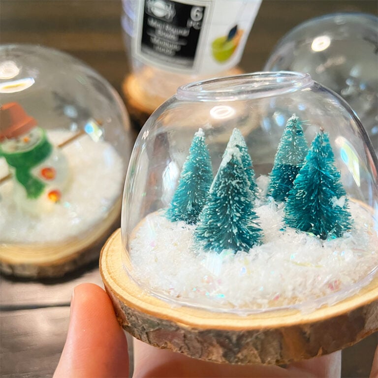 DIY snow globe feature image shows a small snow globe of mini bottle spruce bottle brush trees in a mini bowl dome with a wood slice as a base. Behind this featured globe is one made with a round clear plastic ornament and a wood slice with a 3 inch snowman ornament inside.