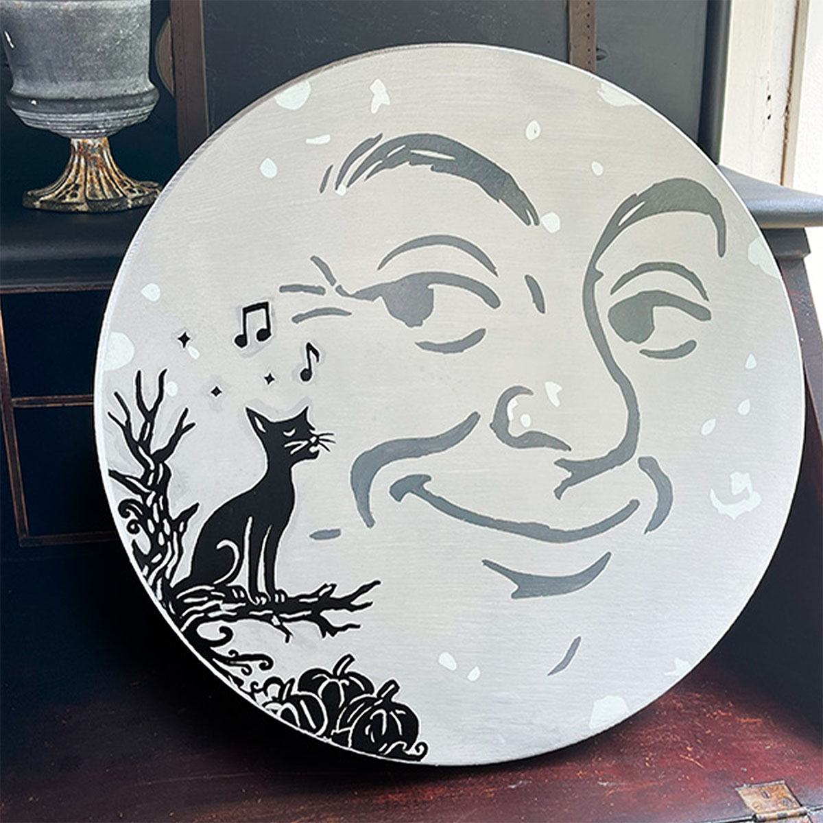 Square feature image of a round man in the moon sign with a signing ally cat on a branch over a pumpkin patch. The sign is gray with darker gray for the moon face and the cat scene is a black silhouette to the left hand side.