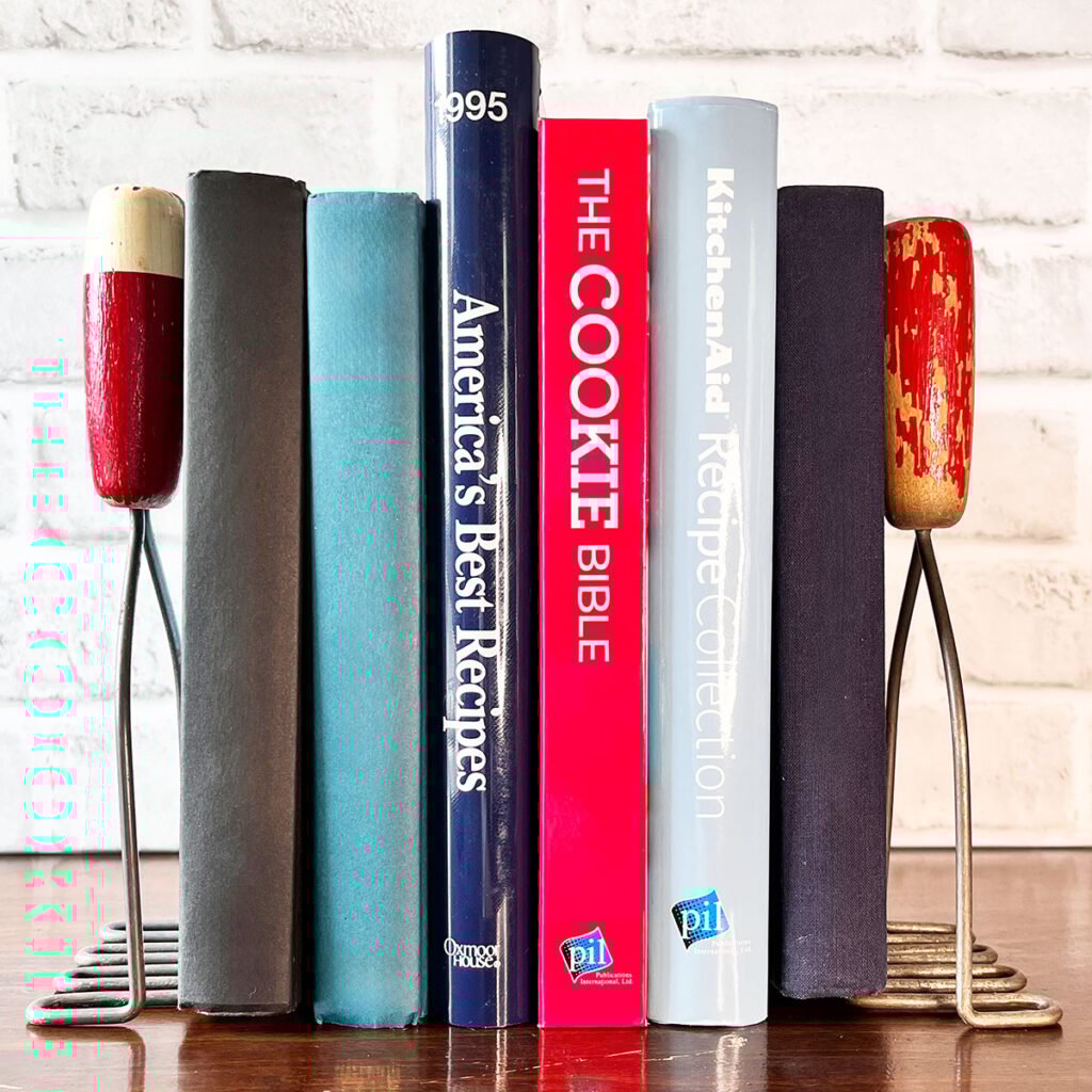Two vintage red handle kitchen mashers used as bookends with blue, red and light blue books in between on a shelf.