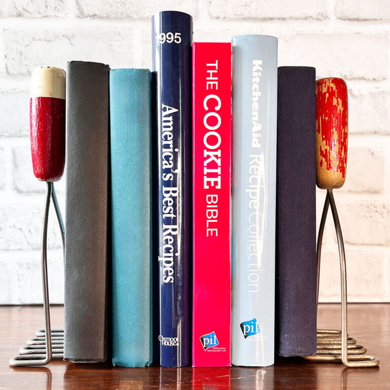 Two vintage red handle kitchen mashers used as bookends with blue, red and light blue books in between on a shelf.