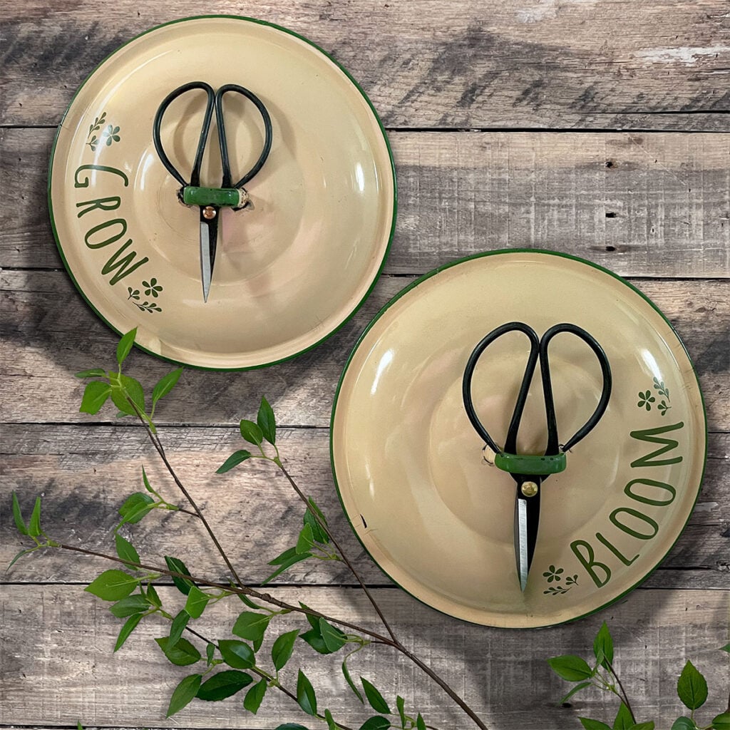 Bloom and Grow feature photo with greenery in the background set on a rustic fir wood wall. Each has a pair of pruning scissors and the art on an arch says 'BLOOM' and the other 'GROW' with tiny flowers. They type and flowers were cut out of olive green cricut permanent vinyl to matched the rim and hand color of the vintage enamel pot lids. The main color of the lids is a warm ivory.