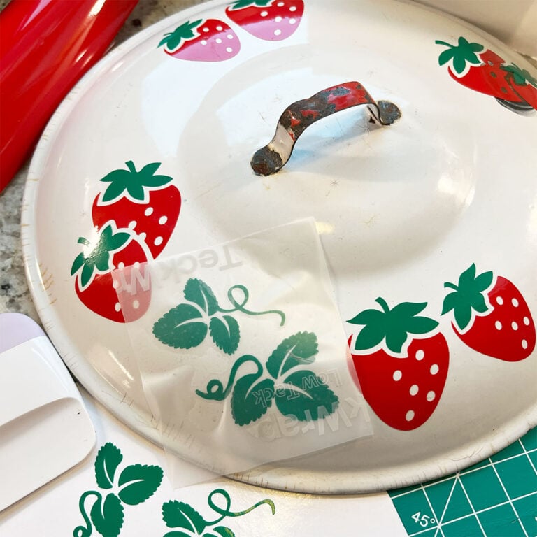 Feature closeup of a vintage enamel white pot lid with a red but rusty handle in the center getting a makeover with beautiful red strawberries with green leafy tops and strawberry leaf vines and greenery around on a work surface. These are permanent vinyl transfers cut on a Cricut. This closeup has a welcoming spring/summer vibe for creating.