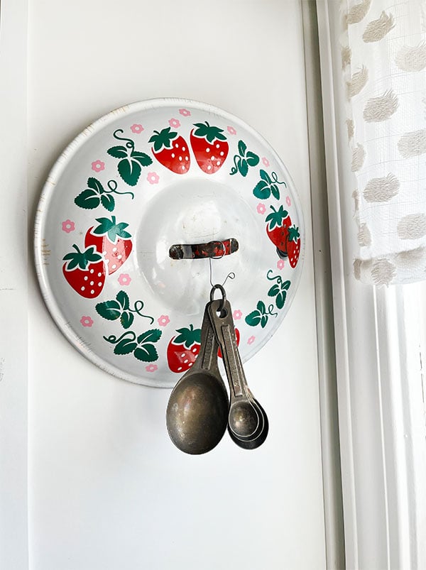 White enamel lid with red strawberries, green leave and vines and small pink blossom flowers pattern around the wide part of the lid. The vintage, chippy enamel white lid that has a rusty c handle in the middle with a touch of it's original red paint. Hanging from a hook on the center handle is a set of vintage aluminum measuring spoons. This cheerful strawberry pot lid hangs on the side of my white kitchen cabinet in the kitchen window with measuring spoons at the ready for baking!