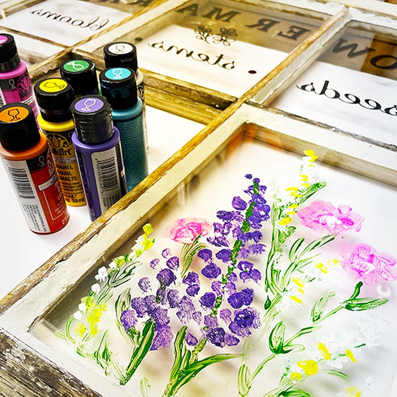Process shot and feature photo for a roundup post of 5 Creative Ways to Repurpose Old Windows showing a set of glass paints on the side with spring flowers painted onto the back of a window paint with type applies in revers on the top three panes of the is six glass pane window. This closeup is for an old window upcycle into Flower Market decoration for spring.