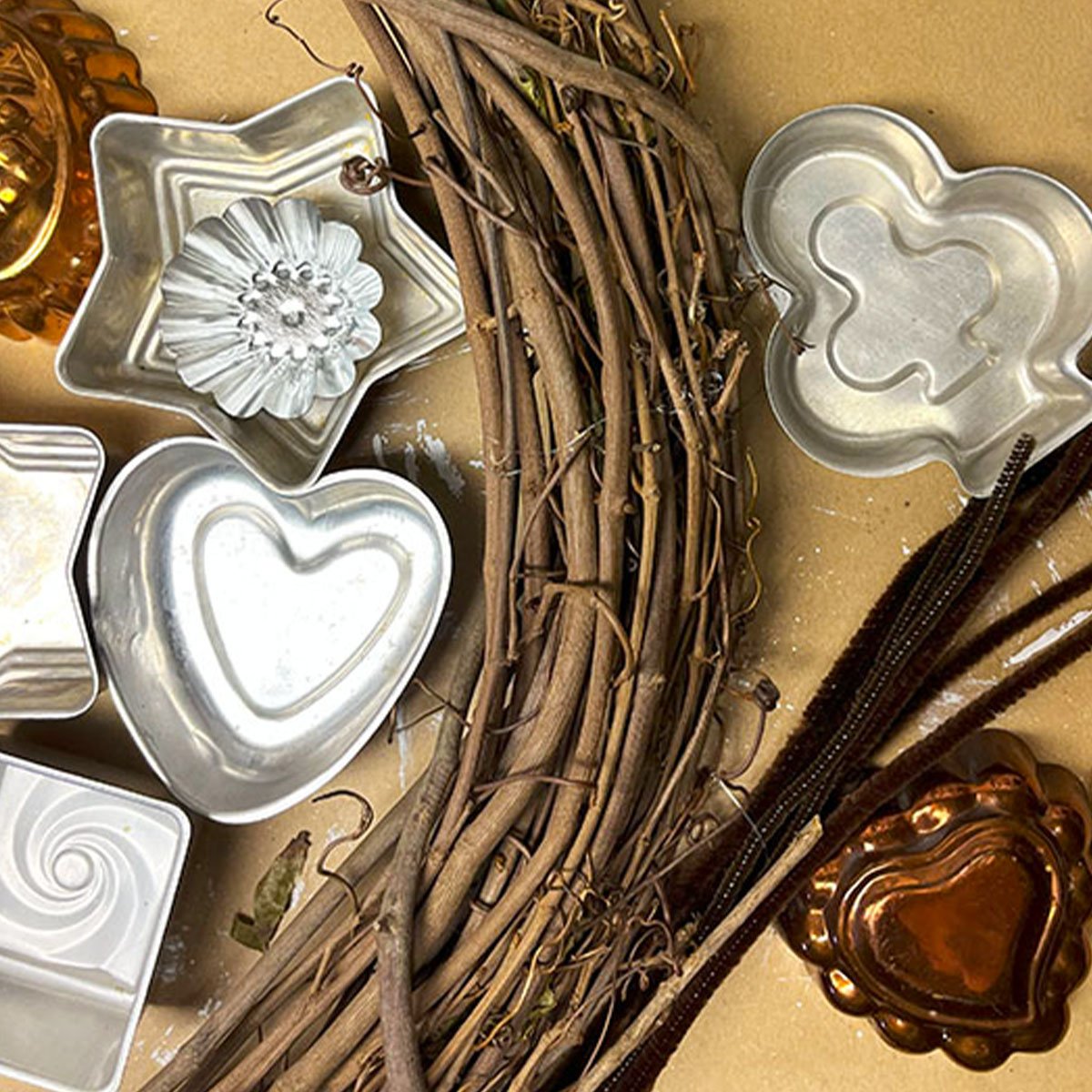 Shown are supplies to assemble a tart tin wreath using a grapevine wreath, an assortment of vintage tart tins (show is a clover, a star, a copper heart, and more.