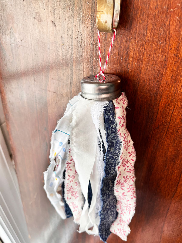 Close up of the metal top to an ordinary salt shake turned into a tassel. The tassel shows scrap fabric ripped into ribbon width in patterned red, blue and white fabrics knotted then secured underneath the metal salt shaker lid.