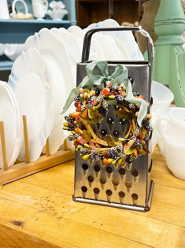Vintage grater given an update with a cute spring mini wreath and beautiful seafoam green wire ribbon at the top of the wreath. The miniature wreath itself has soft white spotted berries in yellow, green, coral and brown.