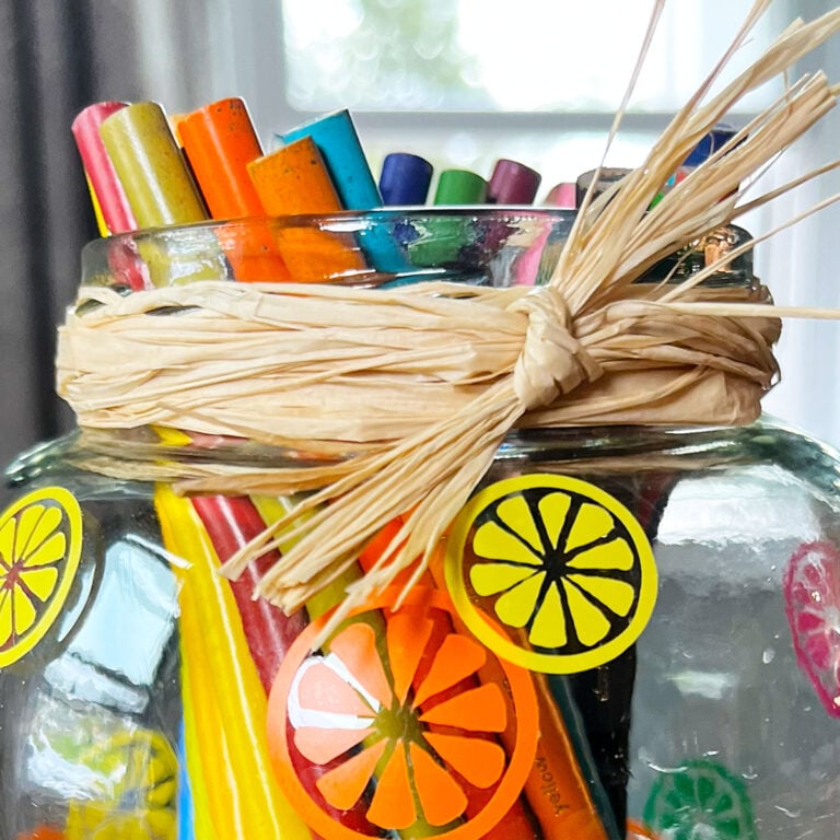 Closeup of the top of a wide mouth jar upcycled into a fun and colorful pencil holder. The jar is full of color pencils with a citrus slice pattern in yellow, orange,, green and magenta on the jar. The top has a wrap of raffia to echo the summer pattern.