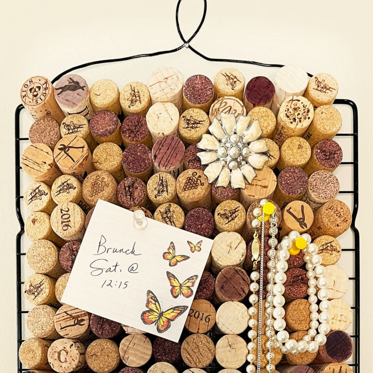 Feature photo of a cork board made from wine corks pushed into a cookie cooling rack sheet. The cork board has a note about brunch with monarch butterflies, a pretty ivory brooch pin and two yellow push pins with pearl bracelets and a necklace hanging from them. A twisted piece of wire (a coat hanger painted black to match) at the top is a decorative element and a practical hanging solution.