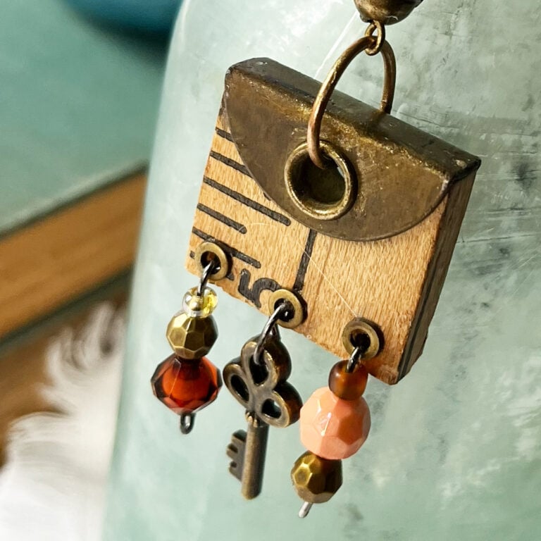 Photo of a metal topped piece from a yardstick with three grommets and jewelry wire hanging from them with peach and amber beads and a tiny vintage skeleton key in the center in brass to match the vintage ruler piece. This closeup shows the beauty of found items, bits and pieces that can be turned from a craft left over to a beautiful statement pendant.