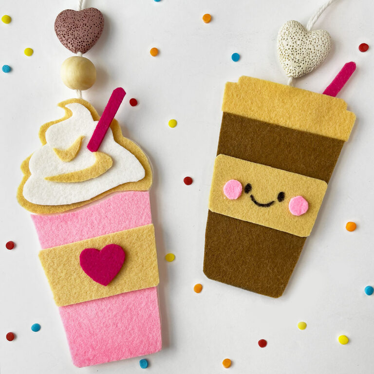 Two to-go style shaped, finished, felt air fresheners shown. The one on the left is a milkshake with a poof of white cream on top and a pink straw with a pick cup and a caramel color wrap with a heart in the center. The cup on the right is a coffee to-go style paper cup with a caramel color lid and center wrap with a smiling face with two pink felt cheeks. Both have lava heart beads at the top in pink and white for the coffee-cup smiling felt air freshener. This is an entirely handmade felt craft that can be scented with your choice of essential oil like peppermint, vanilla or pumpkin spice.