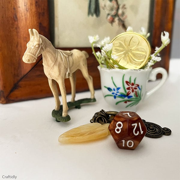 Scene of small trinkets including a vintage horse in ivory plastic cake topper, a small porcelain teacup with floral in red, green and blue with a gold 4-leaf clover and babys breath in the teacup. in front is one multi-sided amber die with 7 at the top and a vintage brass charm with an ivory glass feather.