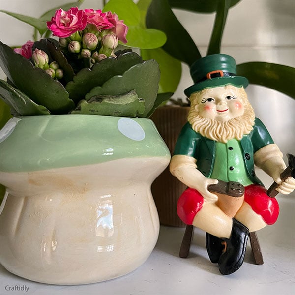 An unexpected vintage leprechaun on a shelf next to a mushroom planter filled with small pink flowers and greenery in the background. The vintage leprechaun has a traditional green hat, jacket, red pants and he is working on re-healing a shoe.