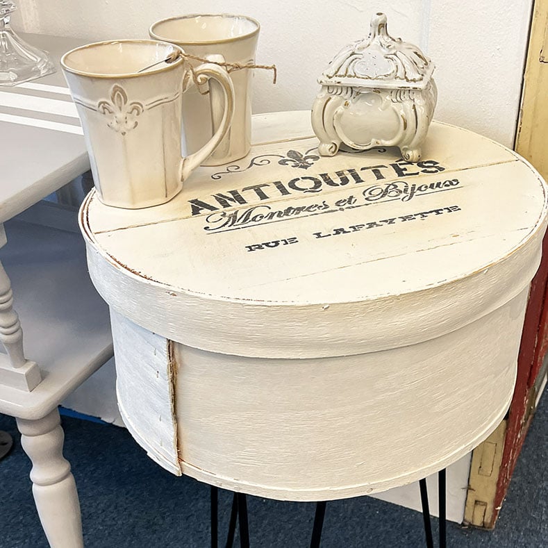 Hatbox turned side table with metal hairpin legs. This feature photo shows the wooden hat box painted white with a stencil: Antiquities Rue Lafayette on it in a flea market booth style with french style mugs and a small ornate ceramic white jewelry box.