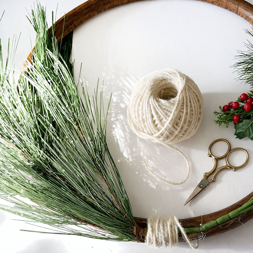In process and photo feature for a roundup of 12 Upcycled Christmas Decor ideas. This wreath is made from thrifted wood ring with greenery, white twine, embroidery scissors and red berry greenery. Here the branch from a fir tree is being tied to the base of the wreath with twine.