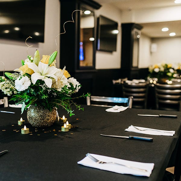 Square photo of the golf course country club room with soft evening lighting that we rented to host a 50th Anniversary celebration for our parents. The room has rich wood tones, ivory colored walls, square tables with dark wood chairs and black tablecloths with beautiful gold and white lily centerpieces in a gold sparkly bubble base with soft tea light (battery opperated) candles and a sparkle of glitter around the floral centerpiece.