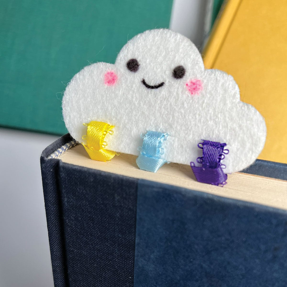 Square feature image of a finished white felt cloud bookmark with three ribbons tied at the bottom in yellow, light blue and purple, sitting at the top of a close book in blue with the cloud at the top and the ribbons holding the place between the book pages.