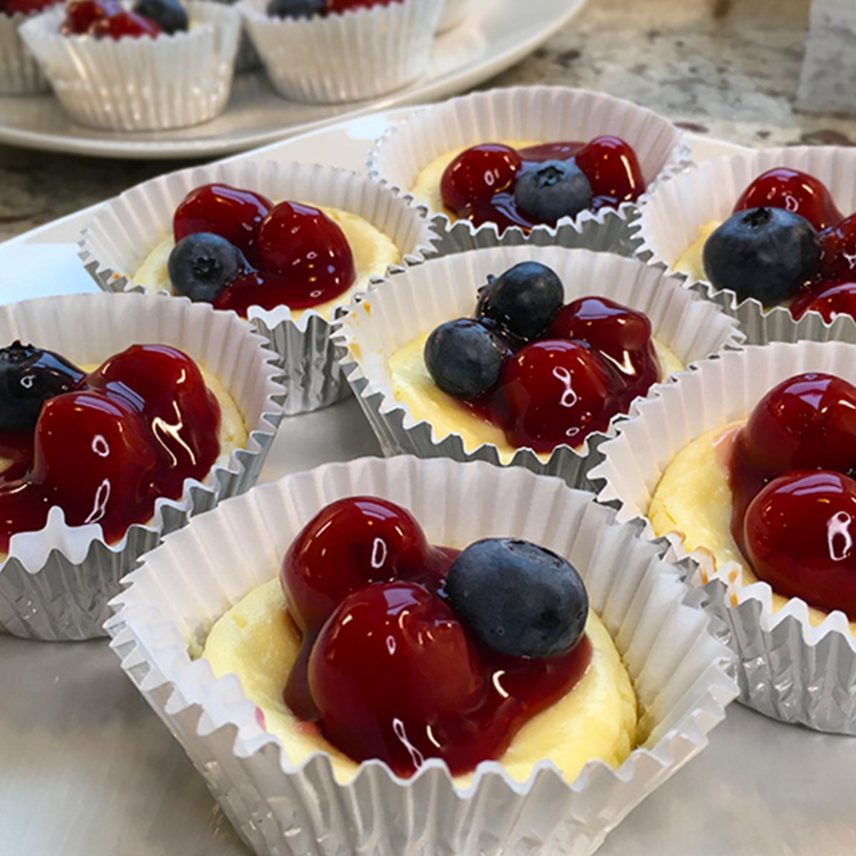 Easy Mini Cheesecakes - this is a square feature image with cupcake liners of individual size cheesecakes topped with cherries in sauce and dotted with blueberries.