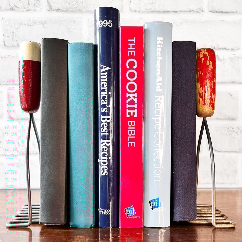 Two vintage red handle kitchen mashers used as bookends with blue, red and light blue books in between on a shelf.