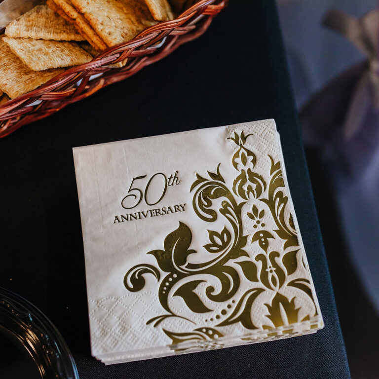 Close up of white cocktail napkins with gold floral print and 50th Anniversary type on an appetizer table with a black tablecloth.