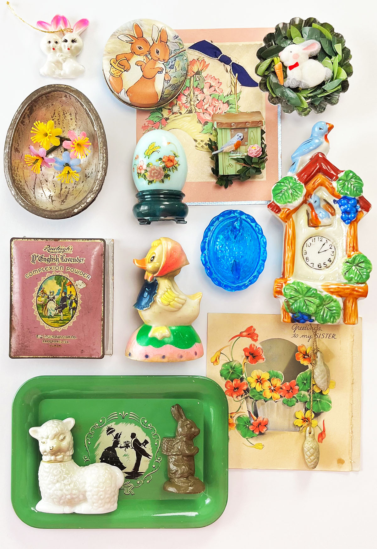 Vintage Easter decor flat lay with small pastel objects arranged in a spring vignette, including a white Avon lamb bottle, pull toy chick, chocolate egg mold, blue glass dish, and handmade rabbit tin magnet in soft blues, pinks, seafoam green, and warm brown tones.