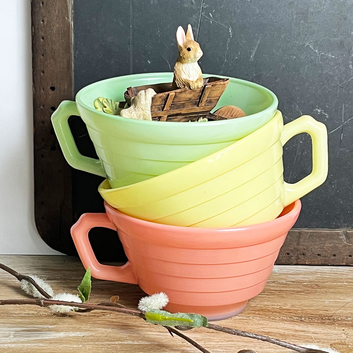 Peter Rabbit figurine in a stack of pastel 1950's vintage teacups against a slate background with warm wood accents. The teacups are peach at the bottom, lemon yellow center and light spring green top. This is a feature image for an Easter Collected post by craftidly with studio art, decor ideas and vintage details. A visual study for the season.