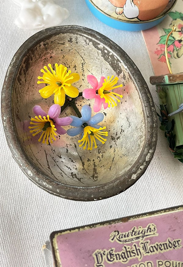 Half of a metal chocolate egg mold with a plastic vintage floral placed inside that is yellow, pink and blue. Nostalgic and fun closeup detail from the large vintage assemblage photo above of Easter and Spring trinkets and treasures from the past shown here in an Easter Collected post and visual study.