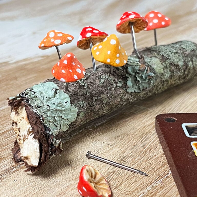 A small stick from outside used as a base for a mushroom scene. The stems of the colorful red, orange and orange-yellow mushrooms are nails. The caps are hand-painted clay with white dot details.