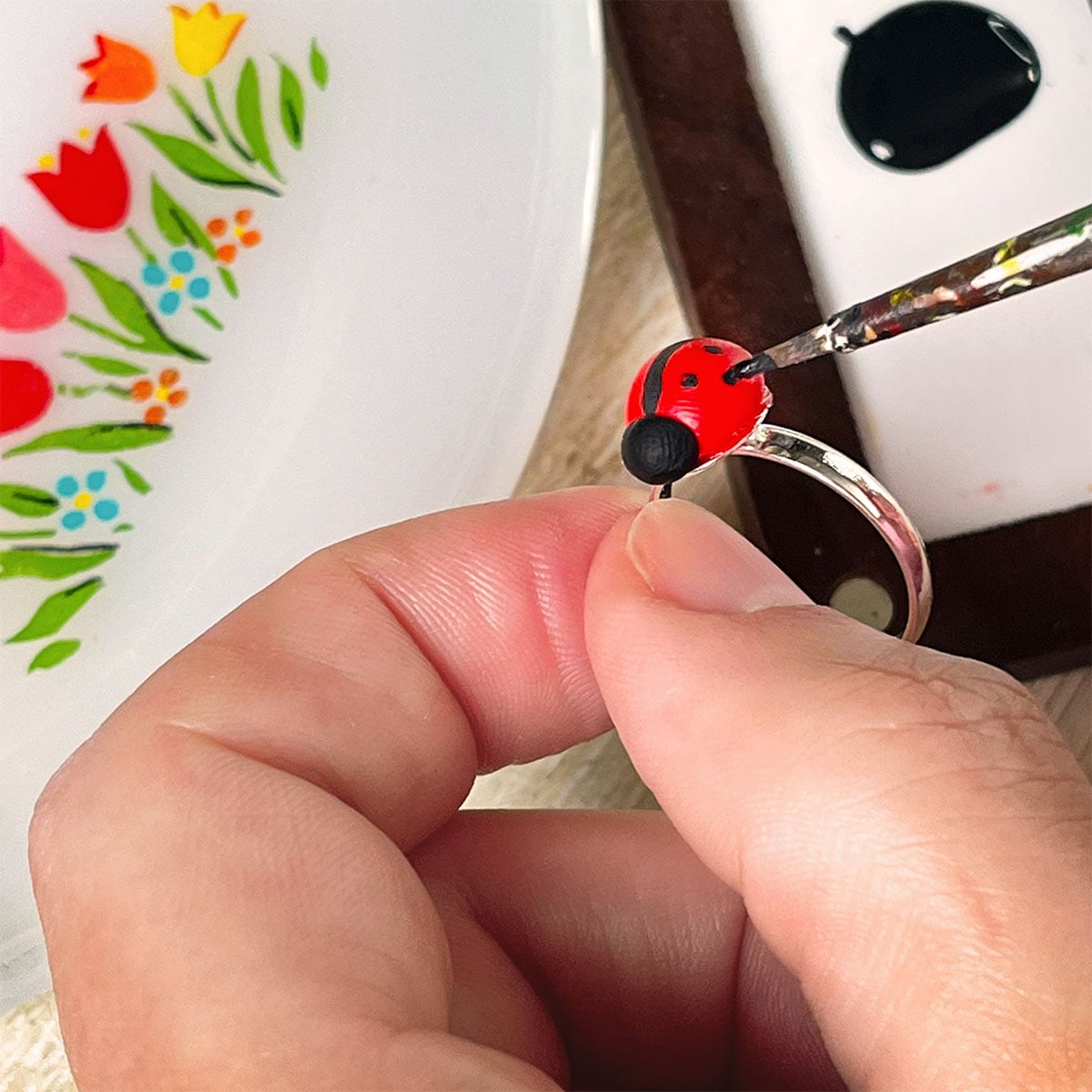 Closeup of a handmade clay red and black ladybug ring being finished with black spots painted on with a fine paintbrush. In the background is a floral vintage milkglass plate with red and yellow tulips on it and a small wood paint palette.