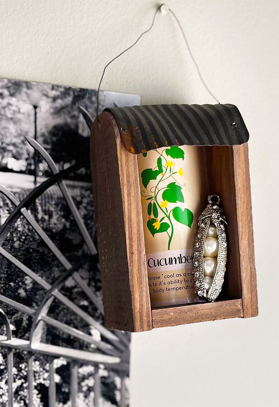Small wood thrifted wall pocket with a tin roof arched over front with an old cucumber playing card and a silver vintage pendant in the shape of peas in a pod. The pod is silver with diamond rhinestones. The peas are pearls in graduating sizes. On the wall next to this piece is a black and white photo of a garden gate.