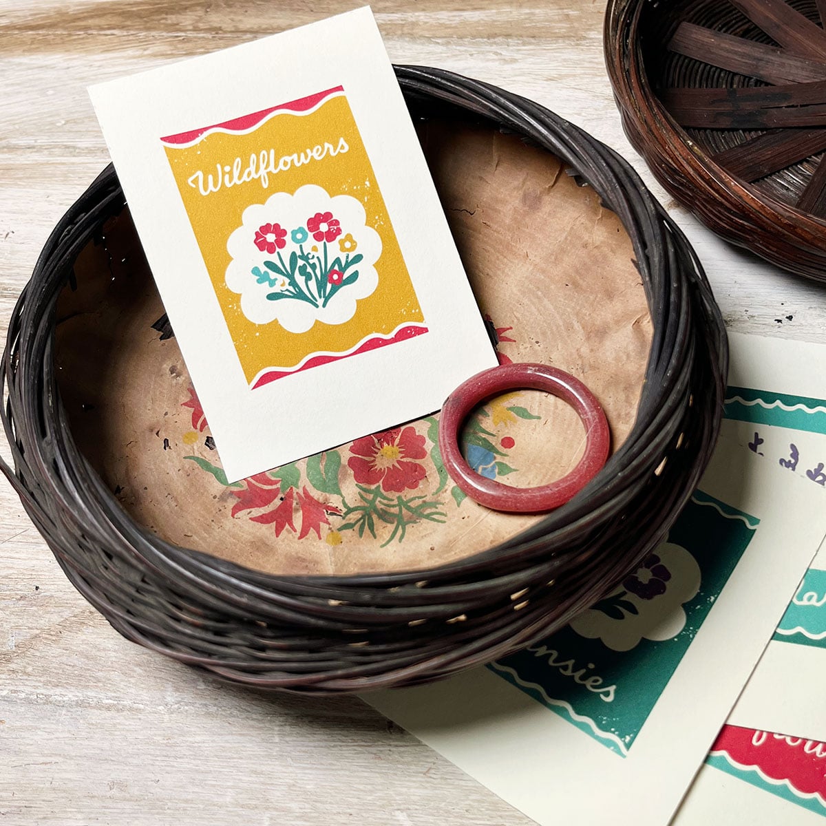 Photo of a wildflower card in vintage yellow, dusty dark pin and pops of deep aqua blue, shot in a vintage round dark wicker sewing basket. The bottom of the basket has a worn old brown paper liner with florals similar to the simple florals of the wildflower card. tucked to the side is the lid of the basket and a glimpse of the other cards in this free printable set.