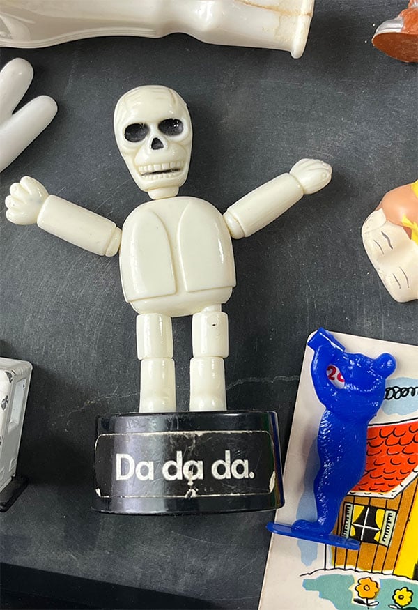 Closeup of a Da da da. skeleton toy held up by elastic and colapses when you push button at the bottom of the base.
