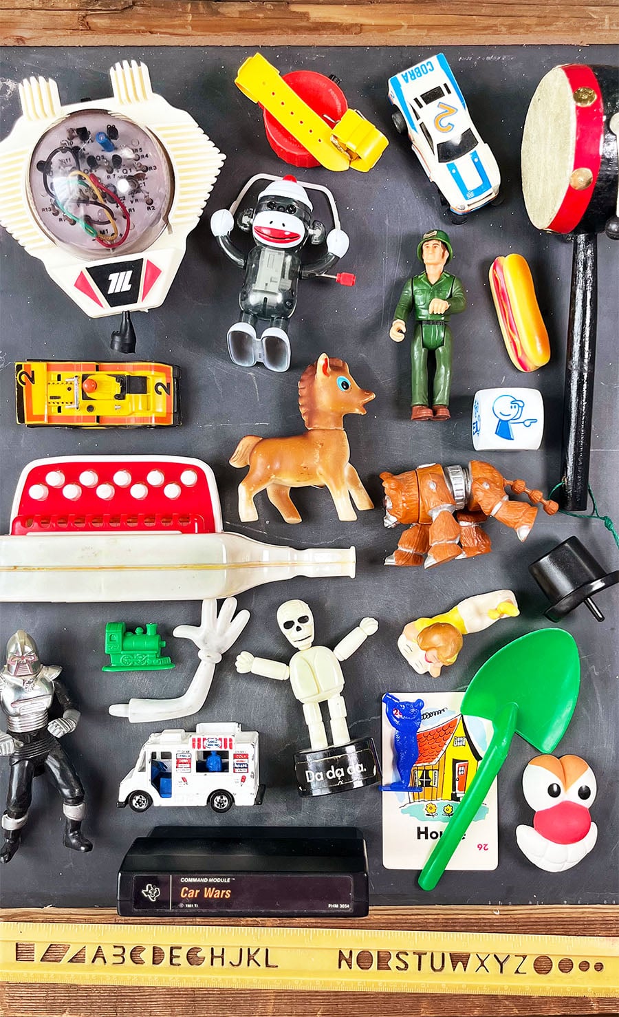 Vintage toys arranged in a flatlay on an old slate chalkboard. There is a rubber squeeker hors, a wind up sock monkey jumping rope, a homemade toy rocket ship, car wars game cassette, hotdog magnet, toy cars and more.