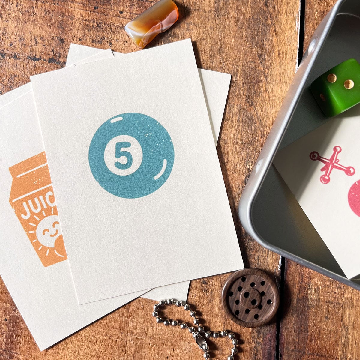 Overhead shot of a que ball in dusty blue on a white cardstock with a smiling sun juice illustration in orange behind it. Next to the cards is a tin with a jack card and green dice, button below. This are free printable keepsake cards with four different illustrations.