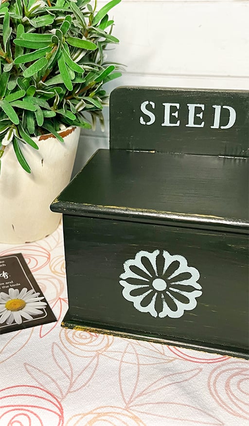 Photo of a wooden seed box made from an upcycled recipe box. The box was painted a chippy olive green. It features a white daisy stenciled on the front and the word 'seed' stenciled in white at the top.