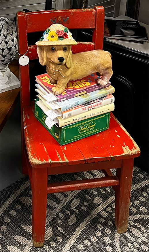 Figurine of a weener dog with a floral bonnet ona stack of vintage childrens books on a child's red wooden chair.