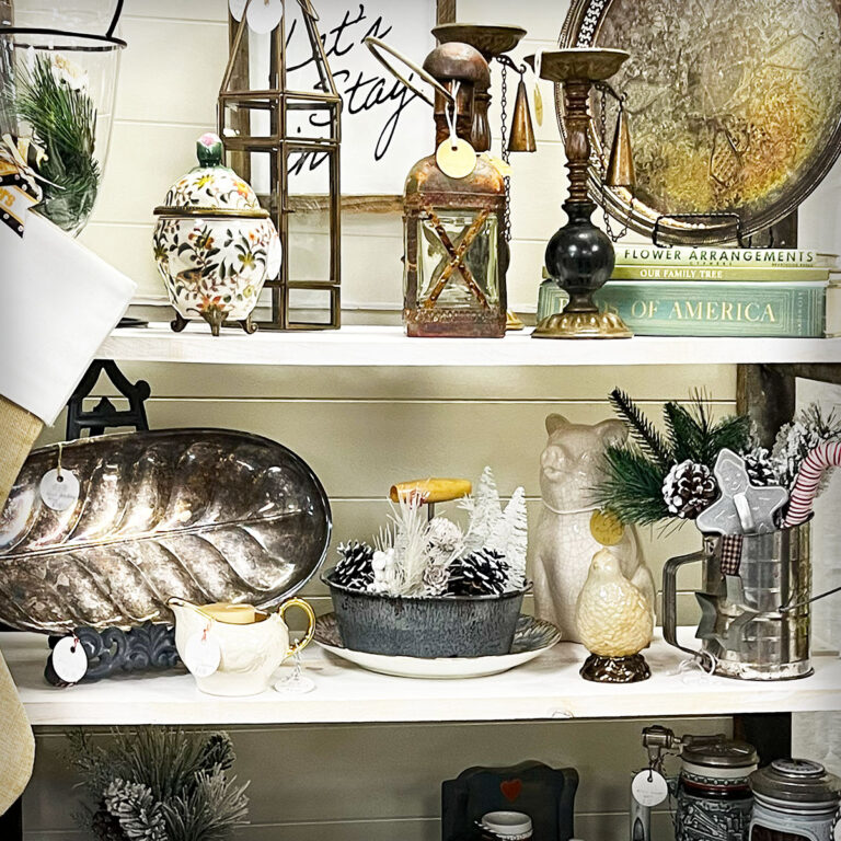Closeup of a vintage booth shelf with wintery items like a brass and glass tall cloche, sterling silver patina pieces in a round and leaf shaped pattern, small vingettes of flocked trees in vintage enamelware, vingtage ooks in green, a quail figuring and a floral porcelain footed jar with lid.