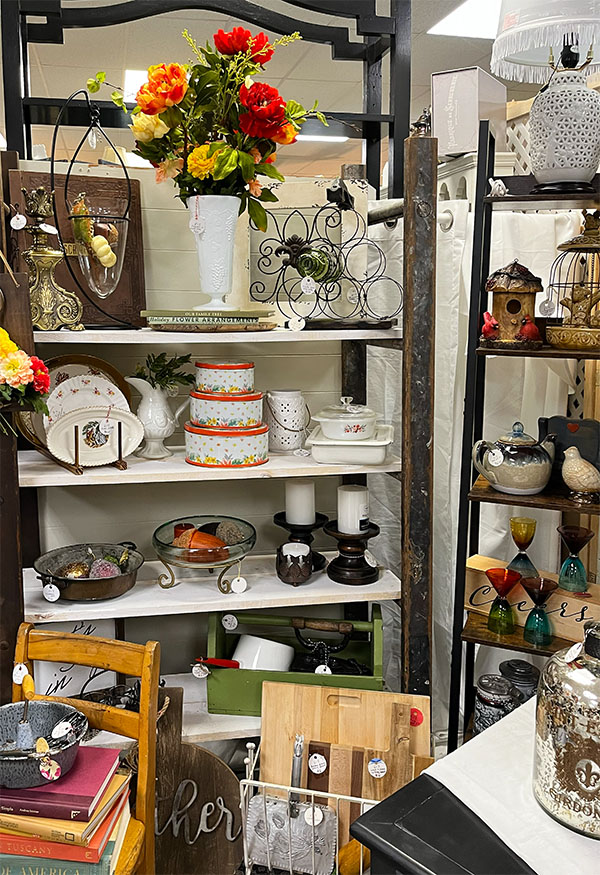 Antique store booth shelf made of old wood ladders filled with Fall vintage and decor including a large vase of orange flowers, orange lidded nesting tins, candlesicks, bird cages, retro glasses and mismatched china plates.