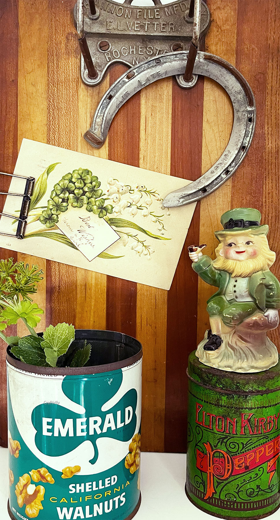 Photo of a vintage clipboard with a lucky horse shoe hanging from the top clipboard hardware below it a brass wire clip holding a vintage post card that has a bouquet of shamrocks. Below that card is a vintage Emerald Shelled Walnuts tin with greenery in it and to the right is a Vintage Elton Kerby Pepper tin shaker in green with red type. Sitting on the large pepper tin is a vintage leprechaun holding a pipe out. His is weathered melamine with a green top hat.