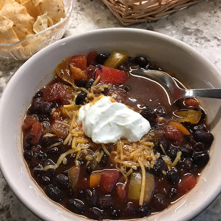 Square feature image of a bowl of black bean soup with onions, tomates, orange peppers, shredded cheddar and a dollop of sour cream on top.