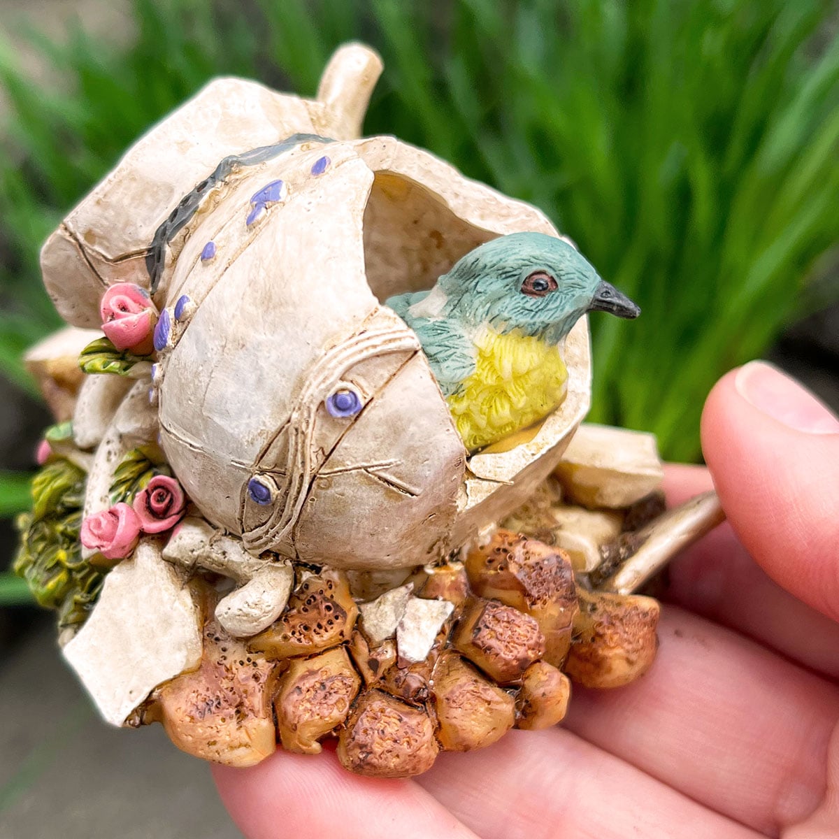 Closeup holding a miniature resin piece that has a decorative broken ceramic jug with a few small roses along the back and a tiny blue bird peering out the hole making himself cozy inside.